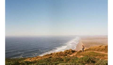 Hiker standing on golden clifftop overlooking a curved coastline with white-capped waves and blue ocean.