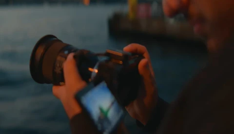 Photographer holding a camera with smartphone mounted below, capturing a sunset over water with boats in the background.