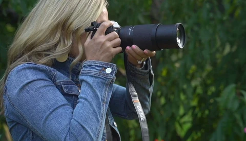 Photographer with blonde hair using a telephoto lens, wearing a denim jacket and sunglasses.