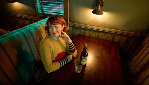 Person with red hair wearing yellow sweater and striped sleeves sits at wooden table with beer bottle under warm and cool mixed lighting.