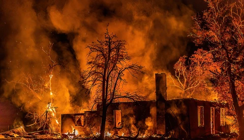 Residential house engulfed in intense flames at night, surrounded by charred trees and heavy smoke.