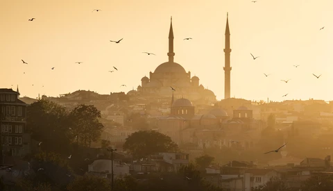 Mosque with dual minarets silhouetted against golden sunset sky with birds in flight.