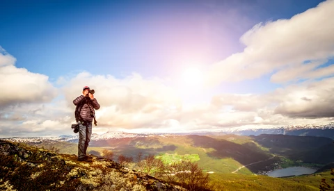 Hiker with backpack stands on a ridge overlooking a vast alpine valley with a lake and snow-capped mountains.