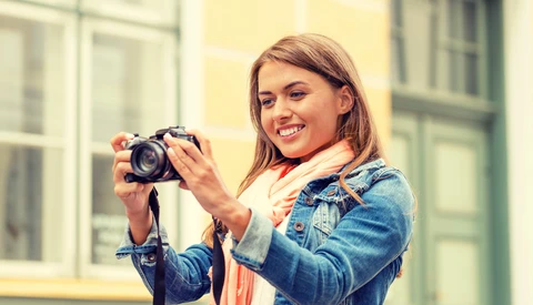 Smiling woman holding a DSLR camera with a telephoto lens, wearing a denim jacket and coral scarf.