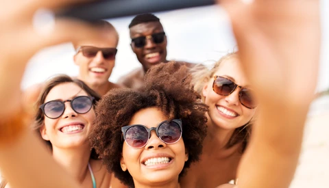 Group of five smiling people wearing sunglasses taking a selfie outdoors in bright daylight.