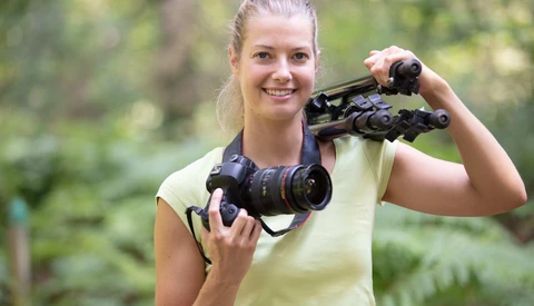 Young woman holding binoculars and a telephoto lens outdoors with blurred green foliage background.