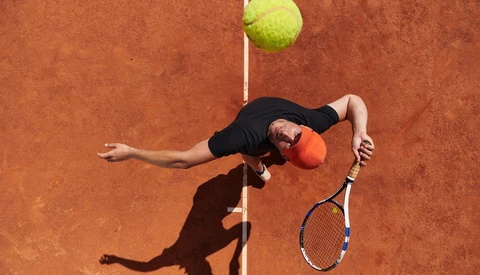 Tennis player executing an overhead serve on a clay court, captured from above with the ball suspended above the net line.