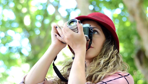 Photographer wearing a burgundy cap and pink shirt looks through a vintage film camera in dappled sunlight.
