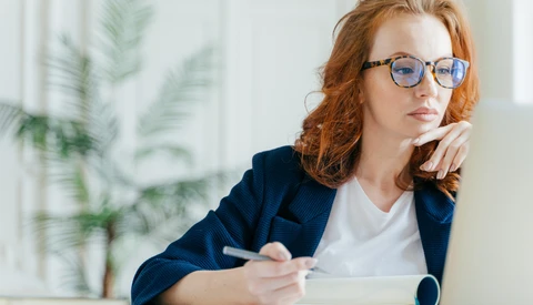 Woman with red hair and glasses working at a laptop while taking notes at a desk.