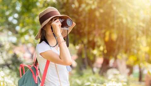Young photographer wearing a hat and sunglasses looking through binoculars in a sunlit outdoor setting.