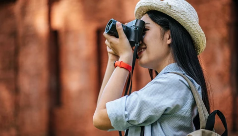 Female photographer wearing a straw hat and light blue shirt holding a camera to her face while photographing against a blurred brick wall background.