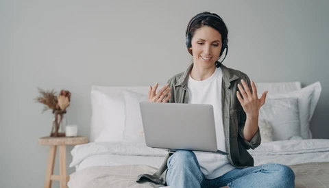 Woman on bed with laptop wearing headset, gesturing while video calling.