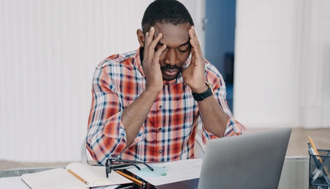 Man in plaid shirt experiencing stress while working at laptop at desk.