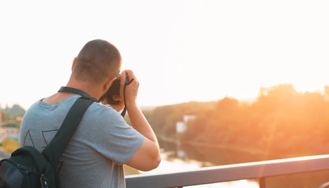 Photographer holding camera to face while standing on bridge at golden hour.
