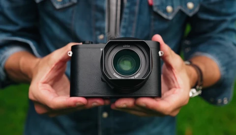 Photographer holding a compact mirrorless camera with a prime lens against a blurred green background.