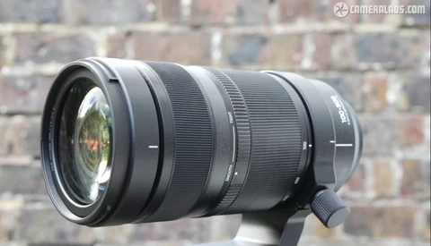 Black telephoto zoom lens mounted on support with brick wall backdrop.