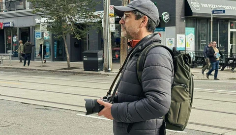 Photographer holding a DSLR camera while wearing a gray cap, black puffer jacket, and olive backpack on an urban street.