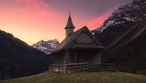 Small wooden church with steeple silhouetted against pink sunset sky and snow-capped mountains.