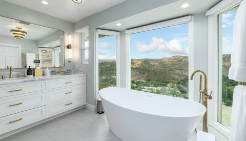 Modern bathroom with freestanding white oval tub positioned in front of large windows overlooking green hillside landscape.