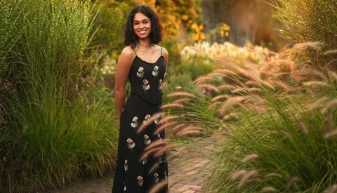 Woman in black floral dress standing among ornamental grasses in golden-hour garden light.