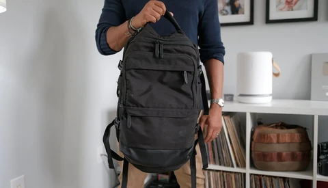 Person holding a black multi-pocket backpack with shoulder straps in an interior setting.