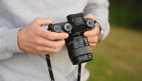 Hands holding a mirrorless camera with a telephoto lens attached, viewed from above against a blurred outdoor background.