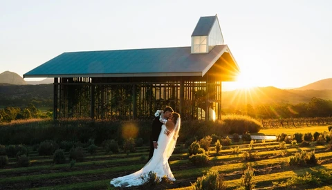 Bride in flowing white gown posing with groom at sunset in front of a modern barn structure with vineyards in the background.