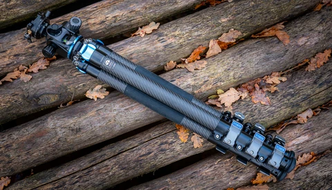 Extended rifle scope with black finish and tactical attachments on weathered wooden logs.
