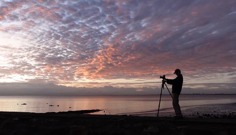 Silhouetted photographer on a beach at sunrise, aiming a camera on a tripod toward the colorful sky.
