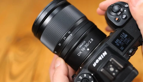 Photographer holding a mirrorless camera with attached zoom lens against warm orange background.