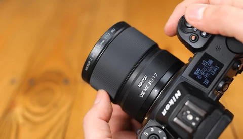 Hand holding a mirrorless camera with a prime lens attached against a warm orange background.