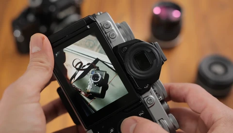 Hands holding a mirrorless camera displaying its rear LCD screen with a self-portrait of the photographer holding the same camera.