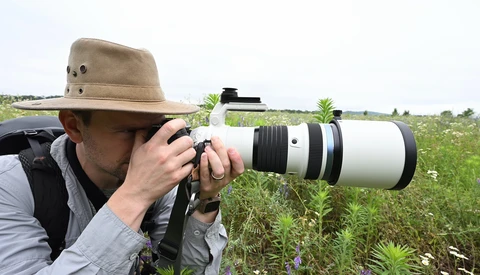 Photographer in tan hat using a white telephoto lens in an overgrown field.