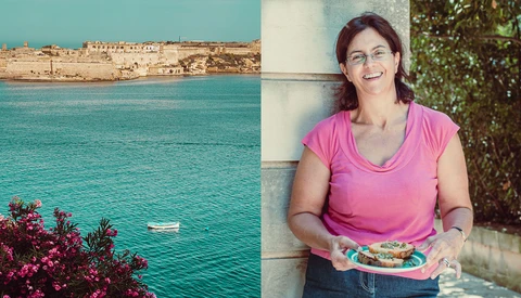 Woman in pink shirt holding colorful food beside Mediterranean coastal landscape.