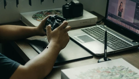 Photographer holding a camera controller while editing video on laptop at desk workspace.
