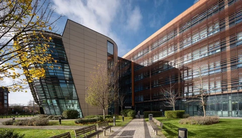 Modern curved glass and metal building beside a brick residential complex with landscaped grounds and wooden benches.