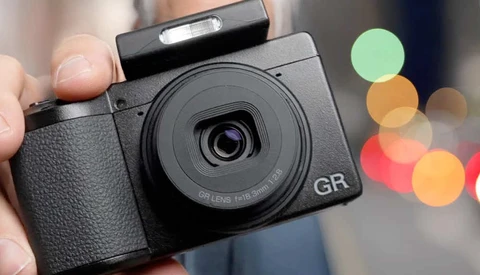 Hand holding a black GR compact camera with a fixed prime lens against a blurred background with colorful bokeh lights.
