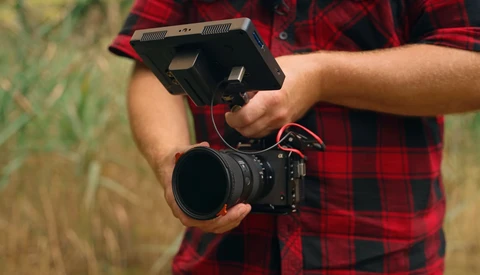 Filmmaker holding a cinema camera with a monitor mounted on top, wearing a red plaid shirt.