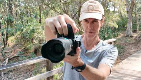 Photographer holding a DSLR with telephoto lens pointed toward camera in a wooded forest setting.