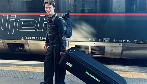 Person in dark formal attire standing beside a black coffin on a train platform next to a passenger train.