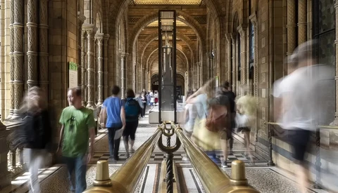 Blurred figures walking through a classical colonnade with arched ceilings and ornate columns.