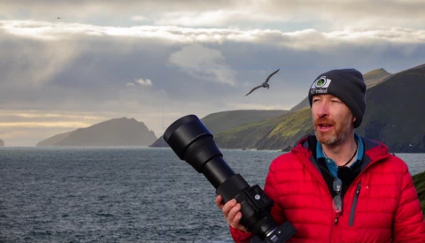 Photographer holding a large telephoto lens while standing on a coastal cliff, with a seabird in flight overhead.