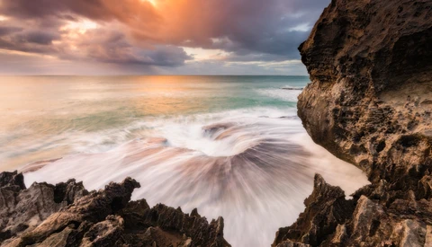 Coastal waves flowing around dark rock formations during golden hour with dramatic cloud-filled sky.