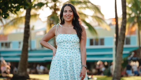 Woman in light blue floral dress standing in park with palm trees and colorful building behind her.