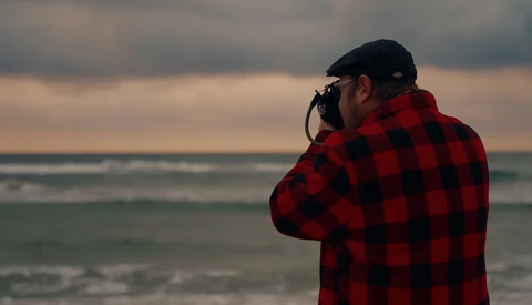 Photographer in red plaid shirt shooting seascape at golden hour from beach.