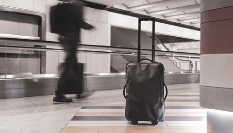 Black rolling luggage sits stationary on a tiled floor while a blurred figure walks past in a modern airport terminal.