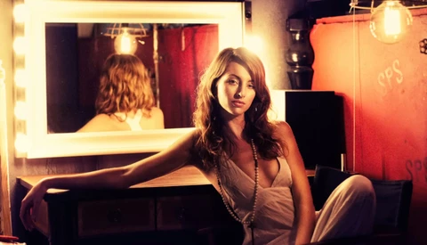 Woman seated at vanity mirror with warm bulb lighting, gazing toward camera in studio setting.