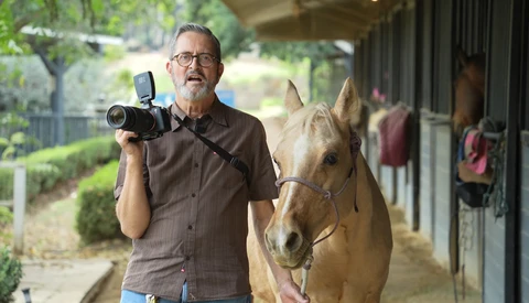 Photographer with vintage film camera beside a brown horse in a stable corridor.