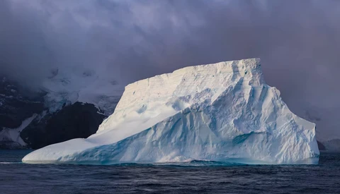 Large tabular iceberg with steep blue-tinted faces floating in dark Antarctic waters.