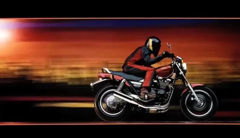 Motorcyclist in red and black leather riding a vintage cruiser motorcycle against a warm sunset gradient background.